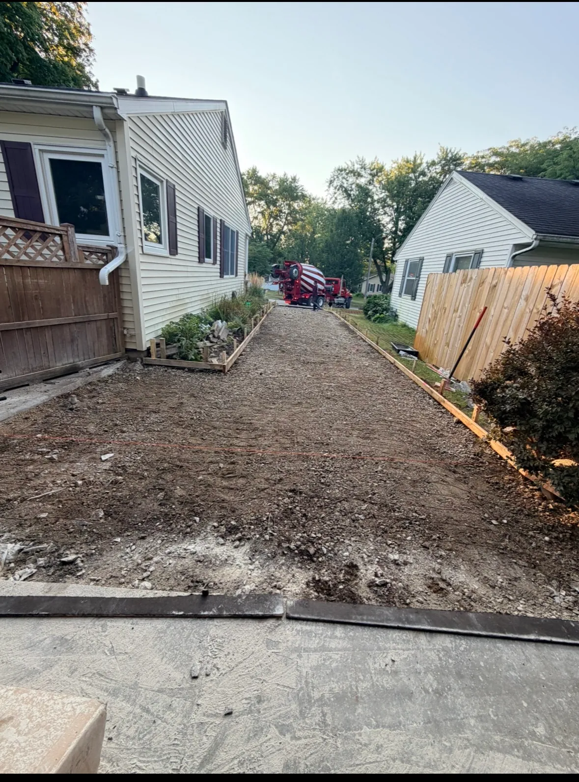 Graded and compacted stone base inside wood forms for a new concrete driveway, with a ready-mix truck waiting at the street