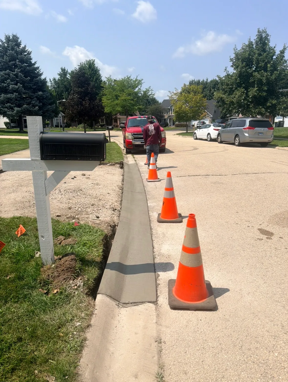 Freshly poured concrete edging along a residential street, coned off to cure, with an Ortiz Concrete Services crew member and work truck beyond