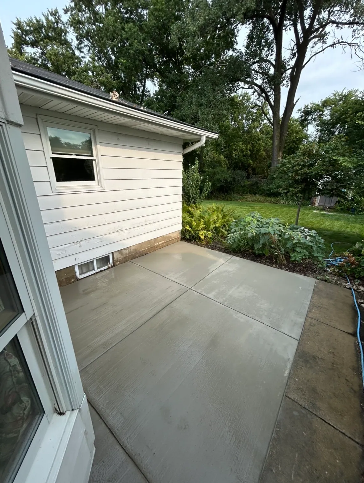 Freshly finished concrete patio slab with tooled control joints beside a white sided home, meeting existing stone paving