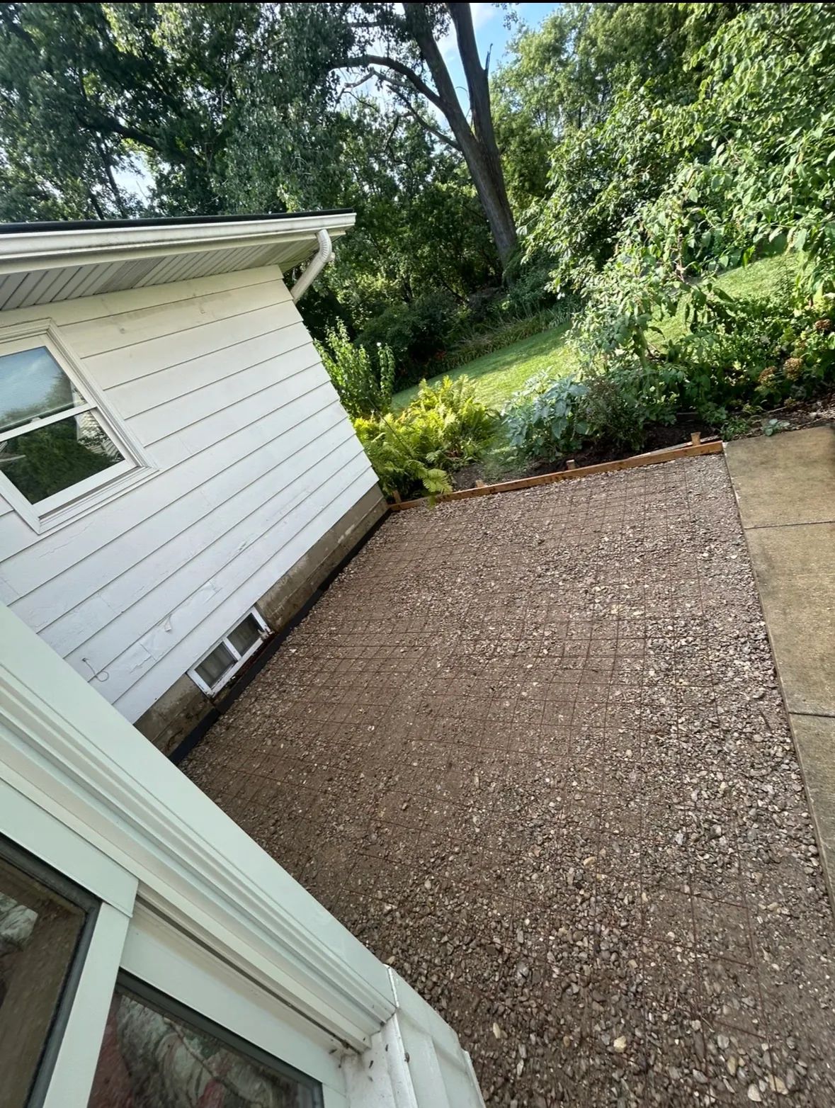 Gravel base with wire mesh reinforcement laid inside wood forms, ready for a new concrete patio pour beside a white sided home